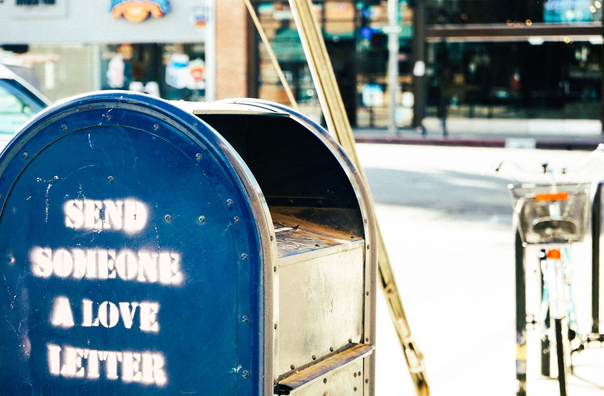A blue mailbox painted with the words Send Someone a Love Letter, on a sunny street.